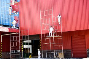 This photo shows a large red commercial building with two sets of scaffolding built against the building. Your professional Pinellas county painters are working on various levels of the scaffolding repainting the exterior of the building. 