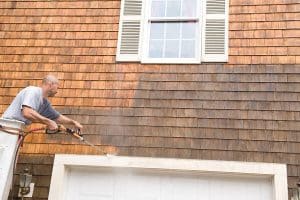 Image is of a worker preparing a surface for professional painting. Man is spraying the exterior of the building with a pressure washer. 