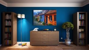 High-quality interior painters near me in Pinellas county showcase a beautifully painted blue wall with bookcases, a potted ficus tree, and a dresser against it. A painting that compliments the wall color is hanging on the wall. 